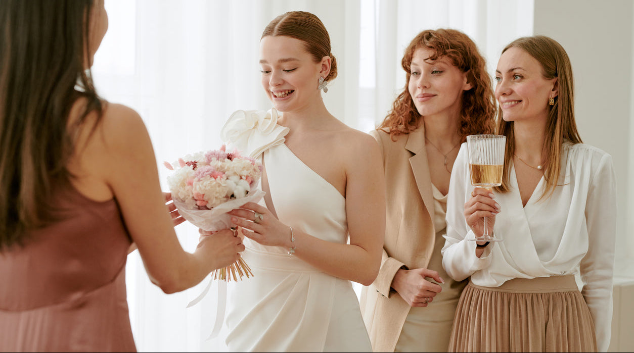 Woman in a white dress receiving flowers from another woman, with two other women watching.